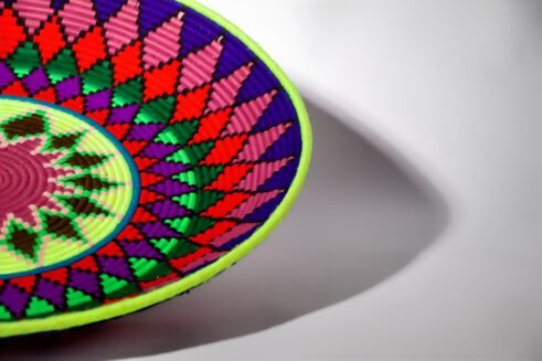 A handcrafted Moroccan coil basket with a vibrant geometric pattern in red, green, blue, purple, and yellow, placed on a neutral background with small stones surrounding it.