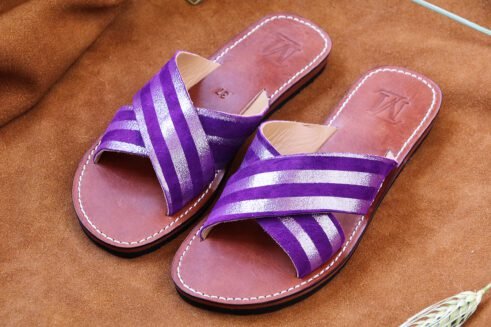 A pair of handmade Moroccan sandals with cross-over striped violet leather straps and brown leather soles, placed on a soft camel suede surface with wheat accents.