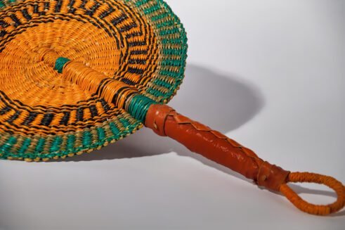 A handcrafted Moroccan fan with a vibrant orange and green woven pattern and a leather handle, placed on a rocky surface with a neutral background.