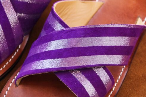 A pair of handmade Moroccan sandals with cross-over striped violet leather straps and brown leather soles, placed on a soft camel suede surface with wheat accents.