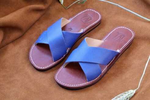A pair of handmade Moroccan sandals featuring vibrant blue leather cross straps, brown stitched leather soles, and a natural beige leather lining, placed on a camel suede background with wheat stems.