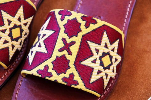 Pair of Moroccan leather sandals with embroidered red and gold star and geometric motifs inspired by traditional zellige tiles, photographed on a camel suede background with wheat stems