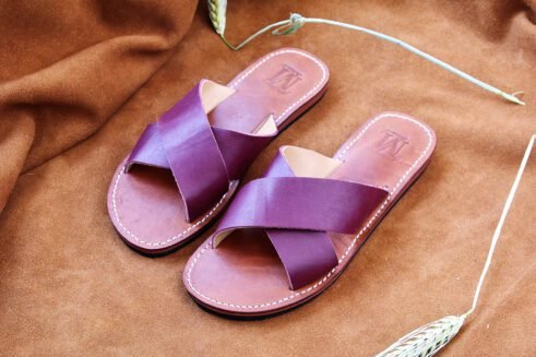 A pair of handmade Moroccan sandals featuring rich plum-colored leather cross straps and brown leather soles with white stitching, styled on a camel suede background with wheat stems