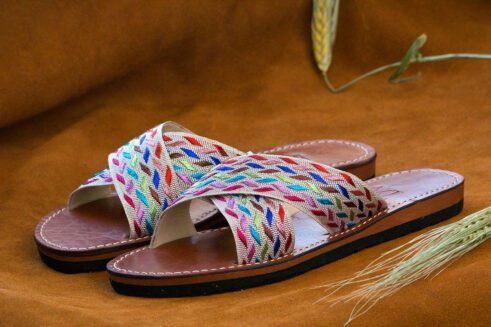 A pair of handmade Moroccan sandals with cross-over braided textile straps in multicolored threads on brown leather soles, placed on a camel suede background with wheat accents