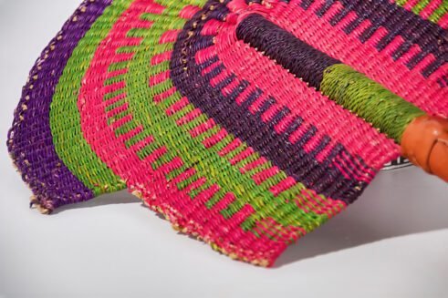 A handcrafted Moroccan fan with vibrant pink, green, and purple colors, complemented by a leather handle, placed on a rocky surface with a scenic background.