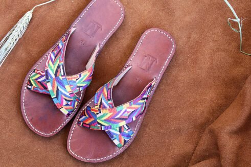 A pair of handmade Moroccan sandals featuring multicolor ethnic-patterned woven fabric in a cross-strap design over brown leather soles, displayed on a camel suede background with wheat accents