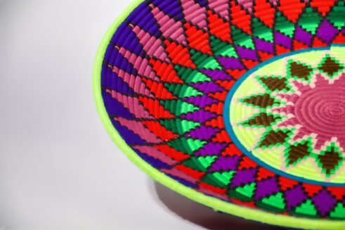A handcrafted Moroccan coil basket with a vibrant geometric pattern in red, green, blue, purple, and yellow, placed on a neutral background with small stones surrounding it.