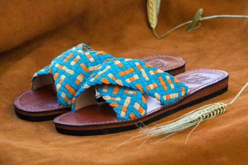 Handwoven leather sandals in blue, orange, and beige tones with a brown sole, placed on a suede background