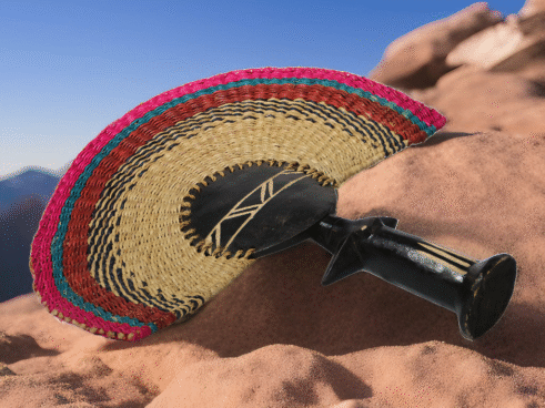 A handcrafted Moroccan fan featuring a colorful woven design with pink, green, and red tones, complemented by a wooden handle, placed on a rocky surface with a clear sky in the background.