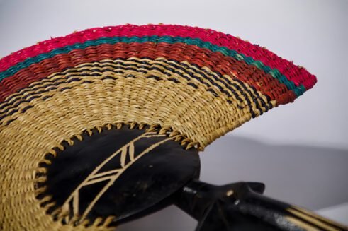 A handcrafted Moroccan fan featuring a colorful woven design with pink, green, and red tones, complemented by a wooden handle, placed on a rocky surface with a clear sky in the background.