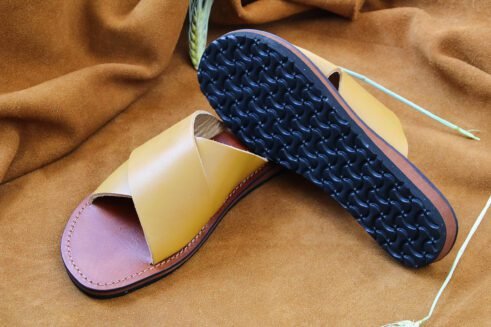 A pair of handmade Moroccan sandals featuring smooth ochre tan leather cross straps, brown leather footbeds, and black rubber soles, placed on a camel suede background with decorative wheat