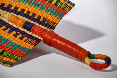 A handcrafted Moroccan fan featuring a vibrant multicolor design with orange, blue, yellow, and red tones, with a leather handle, placed on a rocky surface with a mountain backdrop.