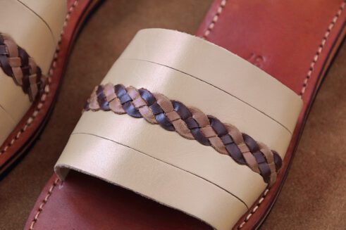 A pair of handmade Moroccan sandals featuring smooth beige leather bands with a dark brown braided detail, brown stitched footbeds, and black rubber soles, styled on a camel suede background