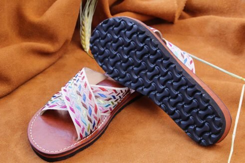 A pair of handmade Moroccan sandals with cross-over braided textile straps in multicolored threads on brown leather soles, placed on a camel suede background with wheat accents