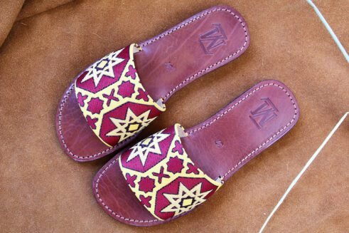 Pair of Moroccan leather sandals with embroidered red and gold star and geometric motifs inspired by traditional zellige tiles, photographed on a camel suede background with wheat stems