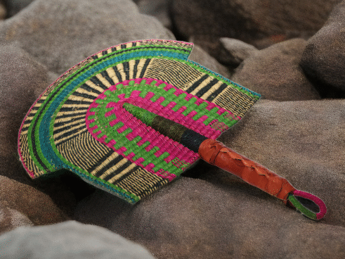 A handcrafted Moroccan fan featuring vibrant green, pink, yellow, and black colors with a leather handle, placed on a rocky surface with a scenic backdrop.