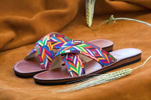 A pair of handmade Moroccan sandals featuring multicolor ethnic-patterned woven fabric in a cross-strap design over brown leather soles, displayed on a camel suede background with wheat accents