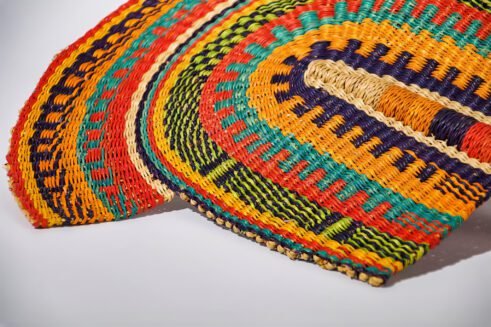 A handcrafted Moroccan fan featuring a vibrant multicolor design with orange, blue, yellow, and red tones, with a leather handle, placed on a rocky surface with a mountain backdrop.