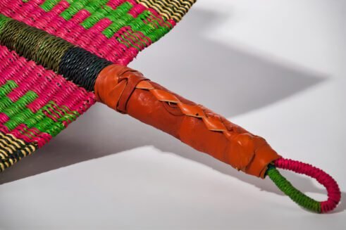A handcrafted Moroccan fan featuring vibrant green, pink, yellow, and black colors with a leather handle, placed on a rocky surface with a scenic backdrop.