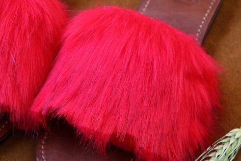 A pair of handmade Moroccan slide sandals featuring vibrant red hair-on leather uppers and brown leather soles, laid on a camel suede background with natural wheat details.