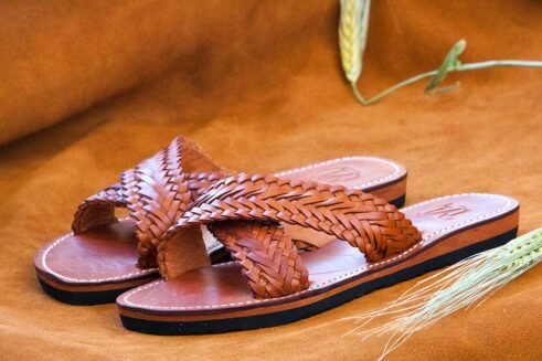 Pair of brown handwoven leather Moroccan sandals with crisscross braided straps, displayed on a warm suede background with barley spikes