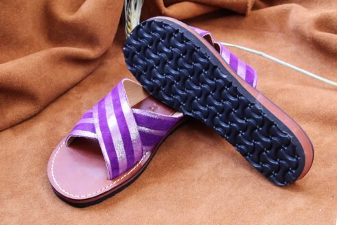 A pair of handmade Moroccan sandals with cross-over striped violet leather straps and brown leather soles, placed on a soft camel suede surface with wheat accents.