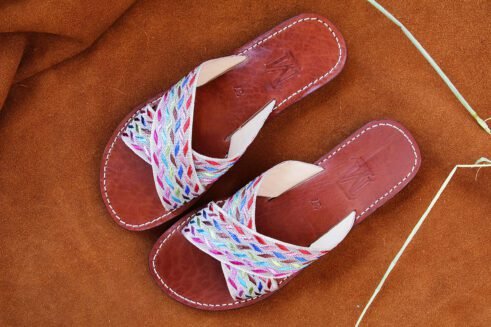 A pair of handmade Moroccan sandals with cross-over braided textile straps in multicolored threads on brown leather soles, placed on a camel suede background with wheat accents