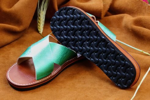 A pair of handmade Moroccan slide sandals with metallic emerald green cross-over leather straps, brown leather soles, and visible white stitching, displayed on a camel suede background.