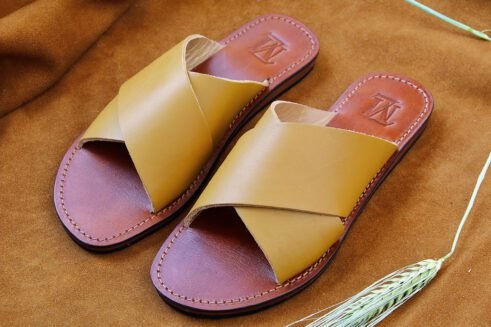 A pair of handmade Moroccan sandals featuring smooth ochre tan leather cross straps, brown leather footbeds, and black rubber soles, placed on a camel suede background with decorative wheat