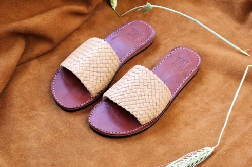 A pair of handmade Moroccan sandals with wide handwoven beige leather straps and stitched brown leather soles, photographed on a camel suede background with wheat accents