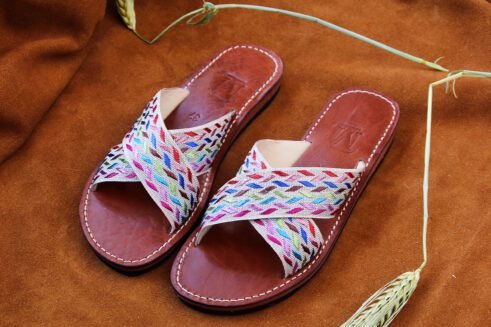 A pair of handmade Moroccan sandals with cross-over braided textile straps in multicolored threads on brown leather soles, placed on a camel suede background with wheat accents