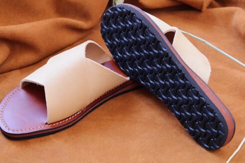 A pair of handmade Moroccan leather slide sandals with beige cross-over straps and brown leather soles, styled on a camel suede background with wheat accents.