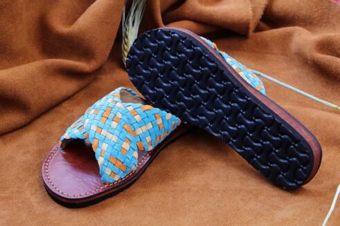 Handwoven leather sandals in blue, orange, and beige tones with a brown sole, placed on a suede background