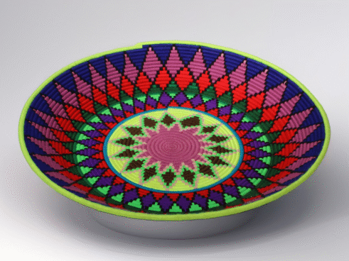 A handcrafted Moroccan coil basket with a vibrant geometric pattern in red, green, blue, purple, and yellow, placed on a neutral background with small stones surrounding it.