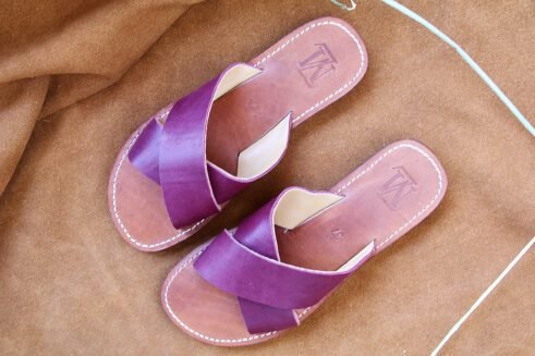 A pair of handmade Moroccan sandals featuring rich plum-colored leather cross straps and brown leather soles with white stitching, styled on a camel suede background with wheat stems