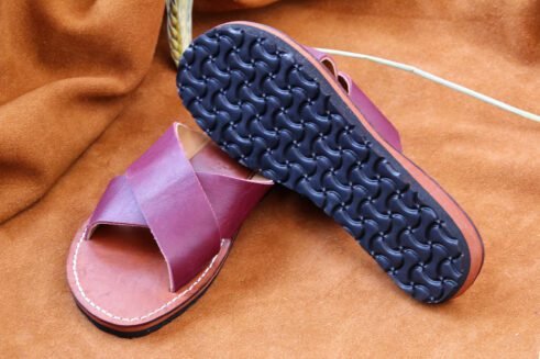 A pair of handmade Moroccan sandals featuring rich plum-colored leather cross straps and brown leather soles with white stitching, styled on a camel suede background with wheat stems