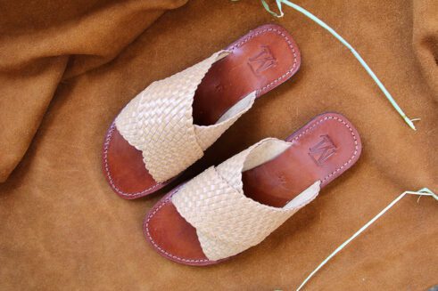 A pair of artisanal Moroccan slide sandals with natural beige handwoven leather straps and dark brown leather soles, displayed on a camel suede fabric with wheat accents.