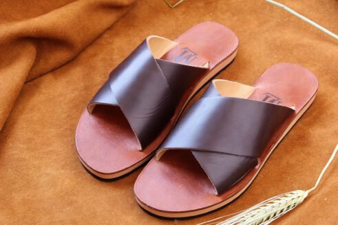 A pair of handmade Moroccan sandals featuring wide dark brown leather cross straps and stacked leather soles with a black rubber base, set on a camel suede background with wheat decor