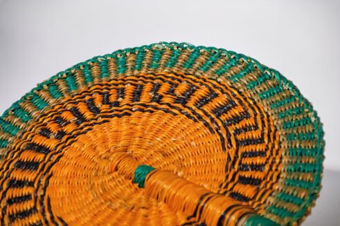 A handcrafted Moroccan fan with a vibrant orange and green woven pattern and a leather handle, placed on a rocky surface with a neutral background.