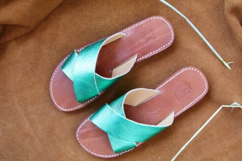 A pair of handmade Moroccan slide sandals with metallic emerald green cross-over leather straps, brown leather soles, and visible white stitching, displayed on a camel suede background.