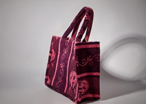 A handcrafted Moroccan tote bag with a floral design in purple and pink, placed on a neutral background surrounded by small stones.