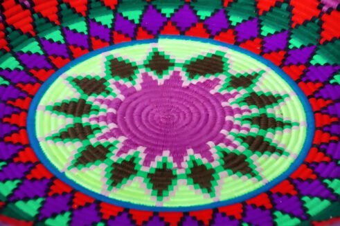 A handcrafted Moroccan coil basket with a vibrant geometric pattern in red, green, blue, purple, and yellow, placed on a neutral background with small stones surrounding it.