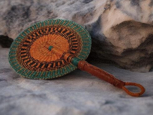A handcrafted Moroccan fan with a vibrant orange and green woven pattern and a leather handle, placed on a rocky surface with a neutral background.