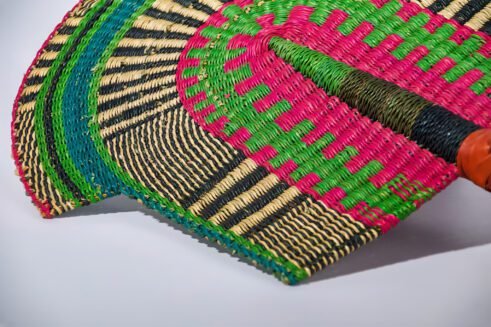 A handcrafted Moroccan fan featuring vibrant green, pink, yellow, and black colors with a leather handle, placed on a rocky surface with a scenic backdrop.
