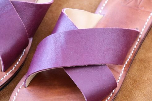 A pair of handmade Moroccan sandals featuring rich plum-colored leather cross straps and brown leather soles with white stitching, styled on a camel suede background with wheat stems