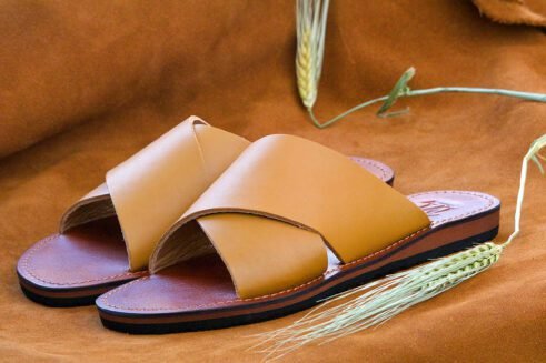 A pair of handmade Moroccan sandals featuring smooth ochre tan leather cross straps, brown leather footbeds, and black rubber soles, placed on a camel suede background with decorative wheat