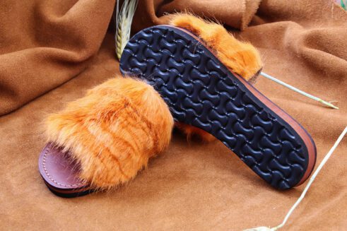 A pair of handmade Moroccan slide sandals featuring a wide burnt orange hair-on leather upper and brown stitched leather footbeds, set against a camel suede background with wheat accents
