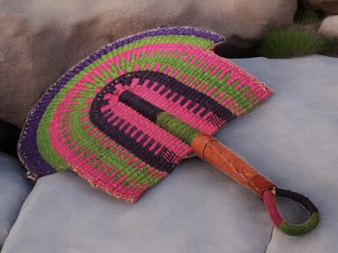 A handcrafted Moroccan fan with vibrant pink, green, and purple colors, complemented by a leather handle, placed on a rocky surface with a scenic background.