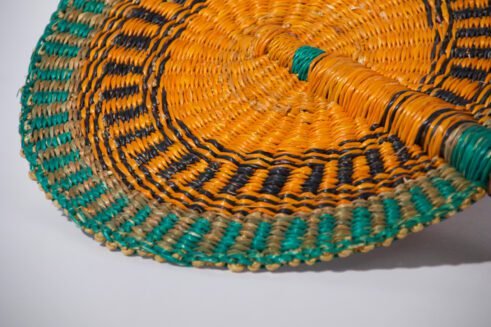 A handcrafted Moroccan fan with a vibrant orange and green woven pattern and a leather handle, placed on a rocky surface with a neutral background.