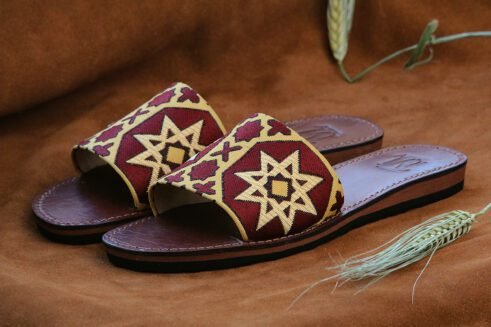Pair of Moroccan leather sandals with embroidered red and gold star and geometric motifs inspired by traditional zellige tiles, photographed on a camel suede background with wheat stems